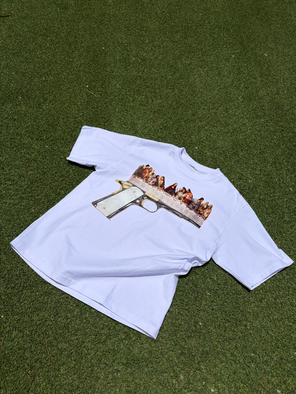 A white short-sleeve t-shirt laid flat on green artificial turf, featuring a graphic print of a handgun superimposed over a painting of the Last Supper.