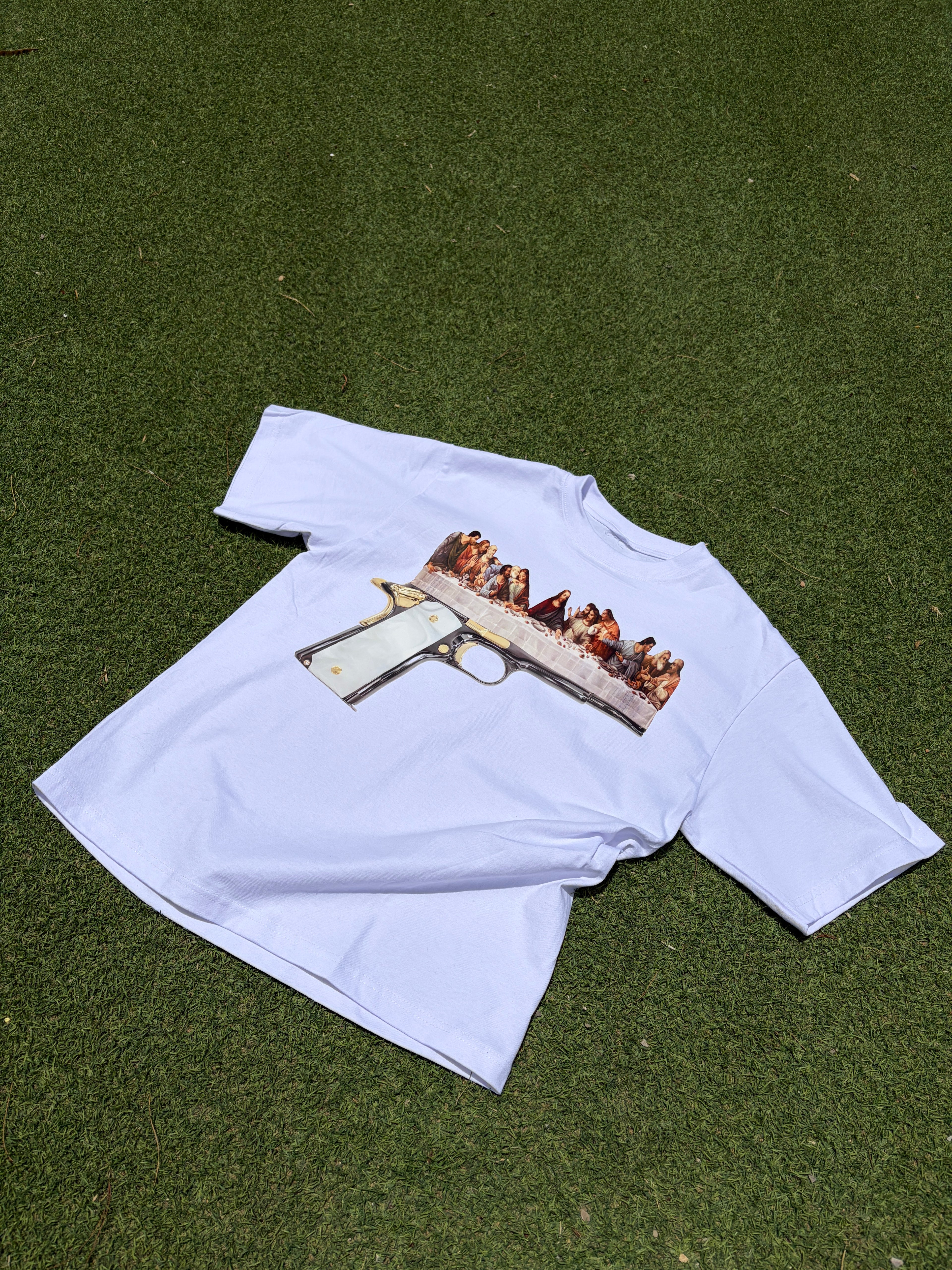 A white short-sleeve t-shirt laid flat on green artificial turf, featuring a graphic print of a handgun superimposed over a painting of the Last Supper.